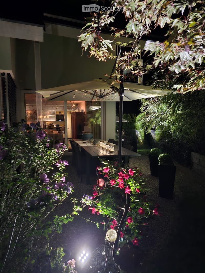 Outdoor patio area with a large umbrella, dining table, chairs, and lush greenery including flowers and potted plants.
