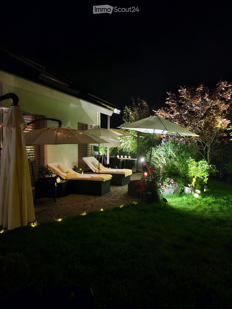 Well-lit outdoor garden area with lounge chairs, umbrellas, and a small table, surrounded by greenery and flowers.