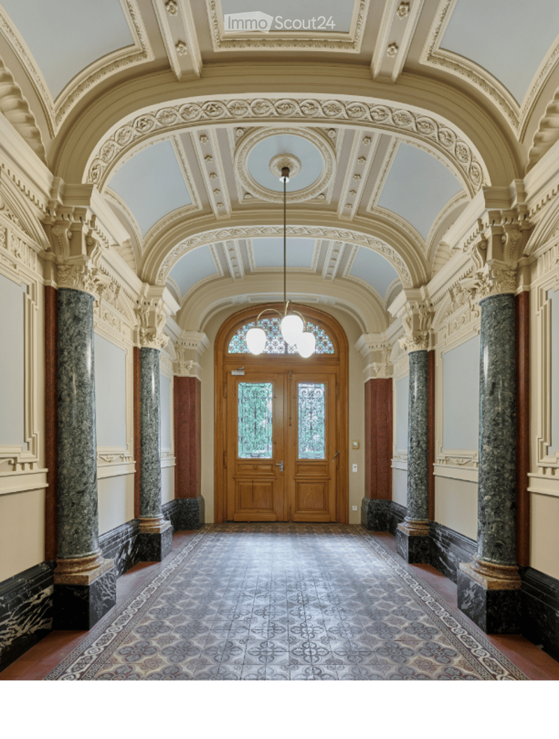 Ornate hallway with high ceilings, decorative moldings, marble columns, and a patterned tile floor. A large wooden double door with stained glass panels is at the end.