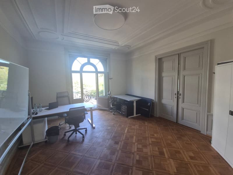 Office space with wooden flooring, large arched window, white walls, and a round ceiling light. Includes a table with chairs, a laptop, a screen, and a cabinet.