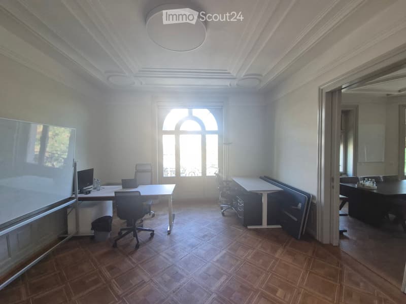 Office space with wooden flooring, large arched window, multiple desks, chairs, and a screen.