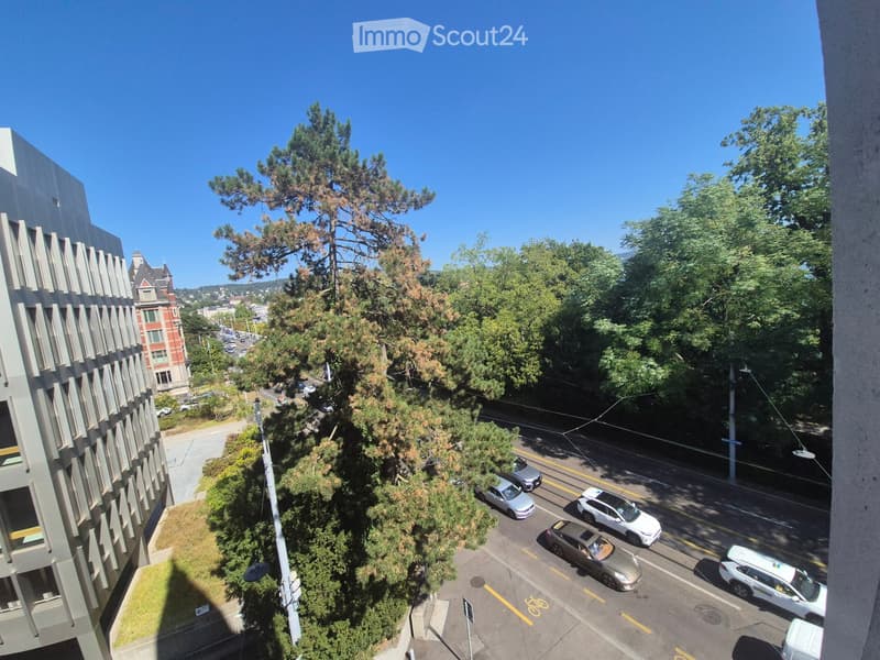A balcony view overlooking a city street with cars, trees, and buildings in the background.