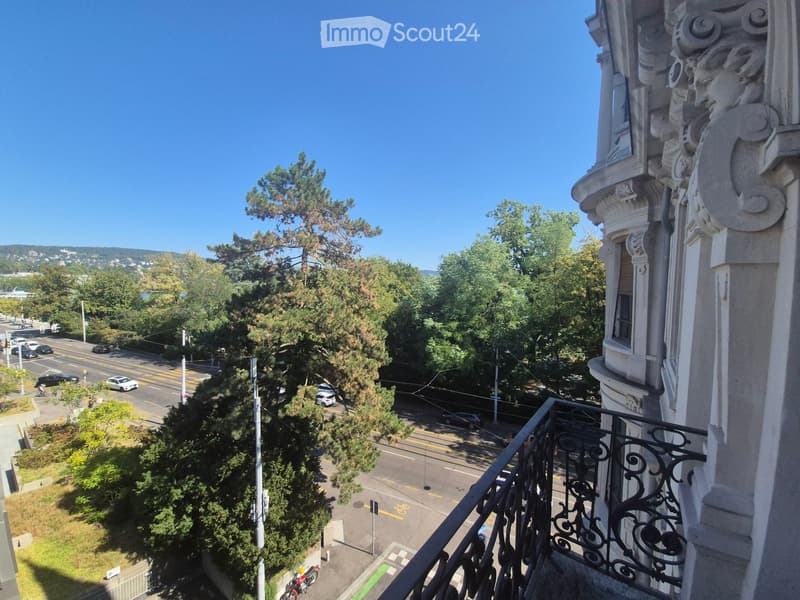 Balcony overlooking a street with trees, road, and distant hills under a clear blue sky.