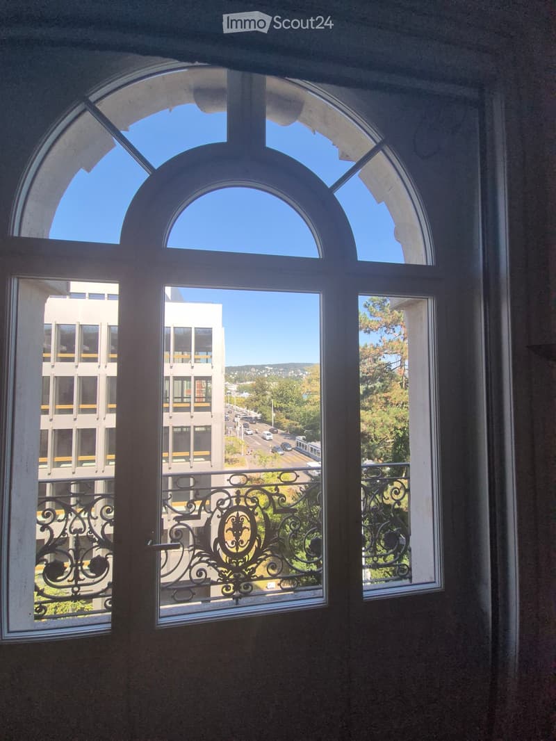 Balcony with ornate metal railing, overlooking a cityscape with modern buildings, trees, and a road with traffic.