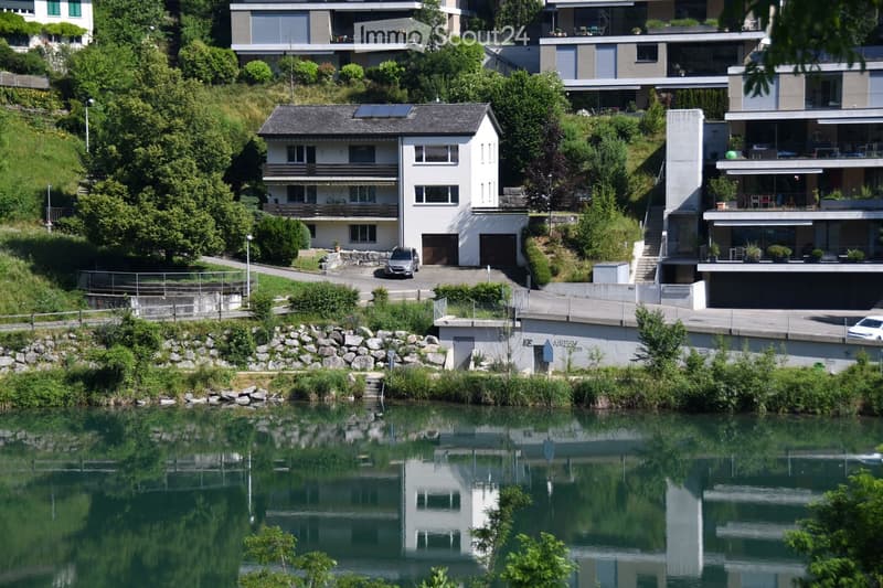 A cluster of modern houses situated on a hillside overlooking a calm river, featuring balconies, large windows, and a mix of contemporary and traditional architectural styles.