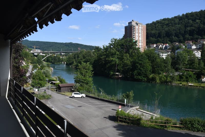 Balcony overlooking a river with a parking area, small shed, and distant buildings and hills.