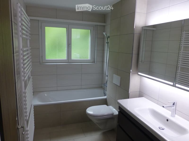 Modern bathroom with a bathtub, toilet, sink, and mirror. Features tiled walls and a window with blinds.