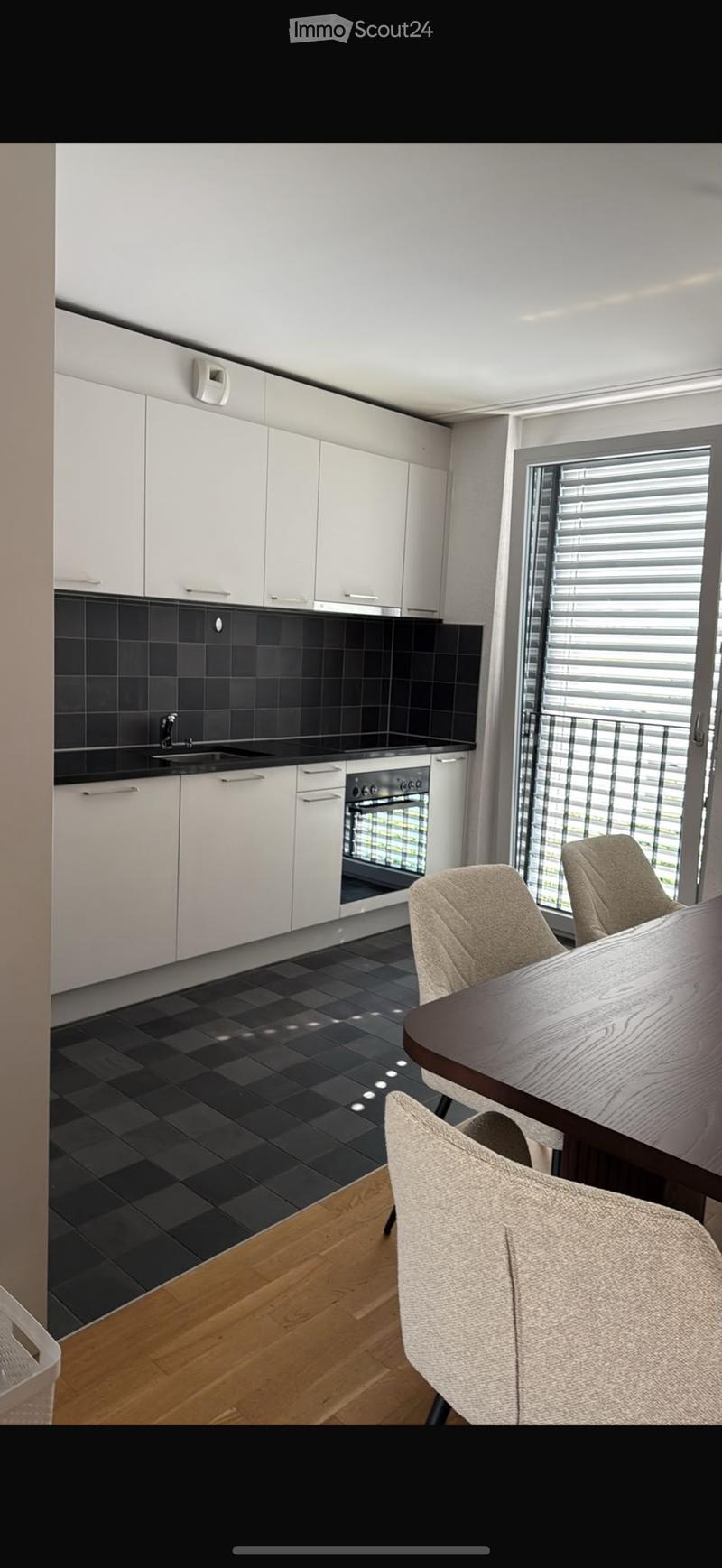 Modern kitchen with white cabinets, black tiled backsplash, and a dining area featuring a wooden table and beige chairs.