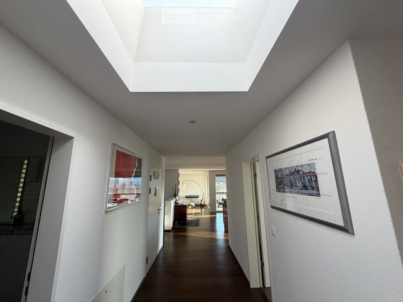 Bright hallway with white walls, wooden floor, and large skylight at the top. Two framed artworks are mounted on the walls, and several doors lead to adjacent rooms.