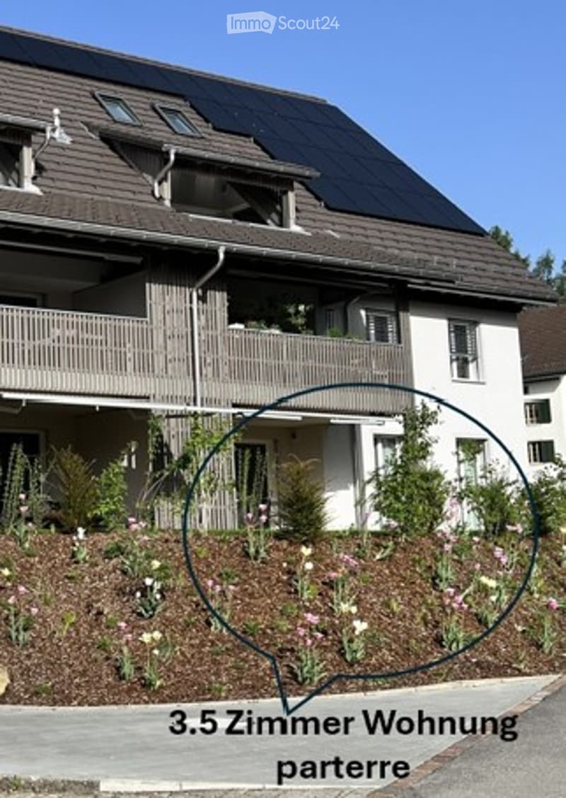Two-story house with solar panels on the roof, white exterior, multiple windows, and a balcony on the upper floor. The front yard features a landscaped garden with various plants and flowers.