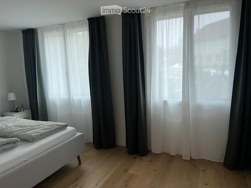 Minimalist bedroom with a single bed, nightstand, and lamp. Large windows with dark curtains allow natural light to fill the space.