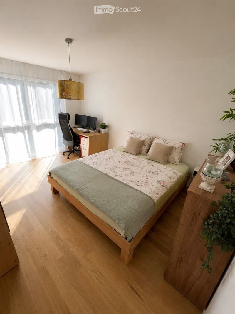 Cozy bedroom with a wooden bed, nightstand, desk with a computer, and a hanging lamp. The room features light wooden flooring and large windows with sheer curtains.