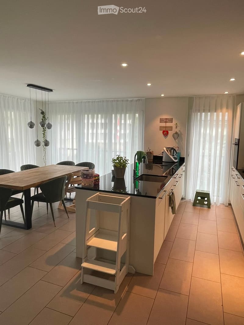 Modern kitchen with a wooden dining table, black chairs, a kitchen island, and pendant lights. The island features a sink, faucet, and a small plant. A green stool is placed near the island.