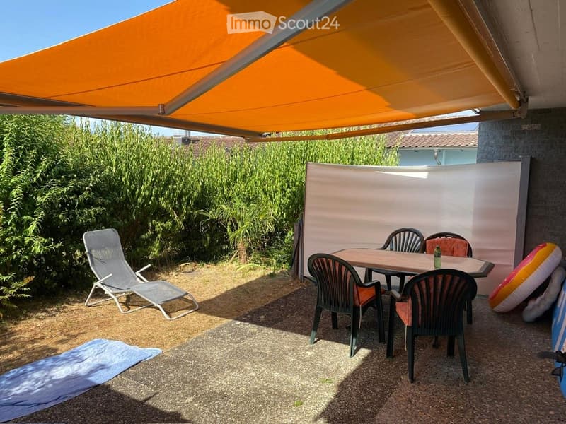 Outdoor patio with a covered awning, a wooden table with chairs, a lounge chair, and an inflatable boat nearby.