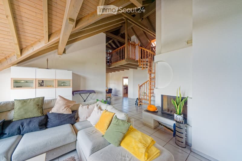 Living room with a grey sectional sofa adorned with colorful pillows, a modern fireplace with a decorative plant, a staircase leading to an upper level, and a white cabinet with glass doors.