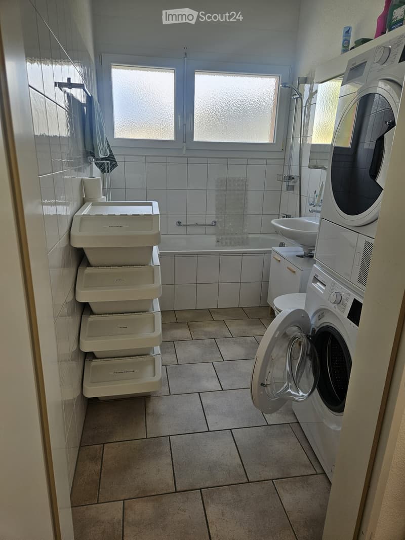 Bathroom with tiled walls and floor, bathtub, sink, toilet, washing machine, and a towel hanging on the wall.