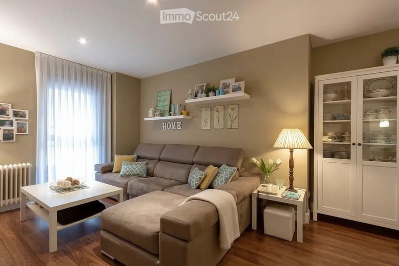 Modern living room with a grey sectional sofa, white coffee table, floor lamp, and decorative cabinet. The room features wooden flooring, large windows with white curtains, and several picture frames on the walls.