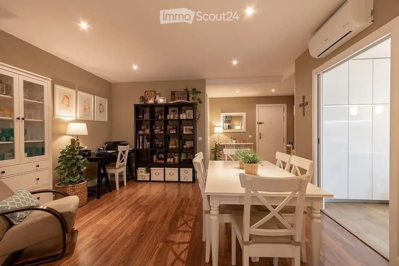 A well-lit, modern living and dining area with wooden flooring, a beige sofa, a white dining table with chairs, a bookshelf, and decorative items.
