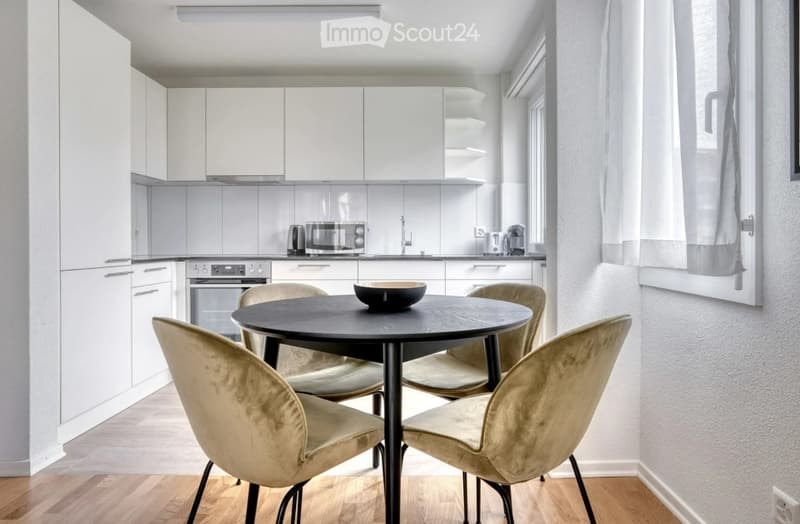 A modern, open-plan kitchen and dining area with white cabinets, a black dining table with four upholstered chairs, and a bowl on the table. The kitchen features a microwave, oven, and sleek countertops.