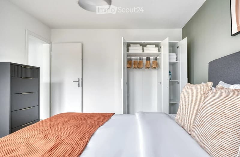 Modern bedroom with a neatly made bed, pillows, a striped blanket, an open wardrobe with hanging clothes, and a grey drawer unit.