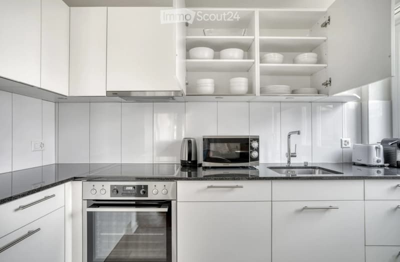 Modern kitchen with white cabinets, stainless steel appliances, and a black countertop. Visible items include an oven, microwave, sink, faucet, toaster, and dishes in the cabinets.