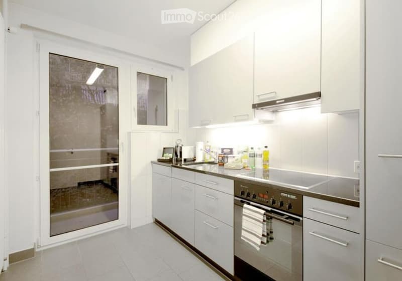 Modern kitchen with white cabinets, stainless steel appliances, and a large window. The countertop features an oven, a faucet, and various cleaning supplies.
