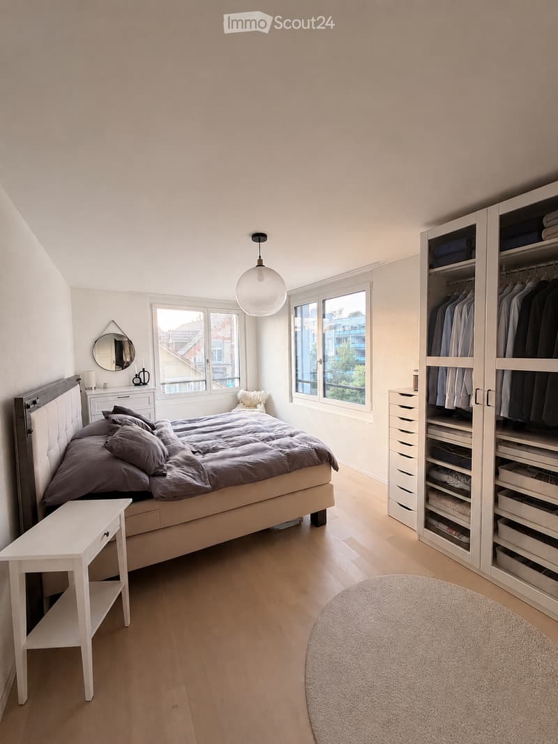 Modern bedroom with a large bed, nightstand, wardrobe, and round rug. The room features wooden flooring, large windows, and a hanging pendant light.
