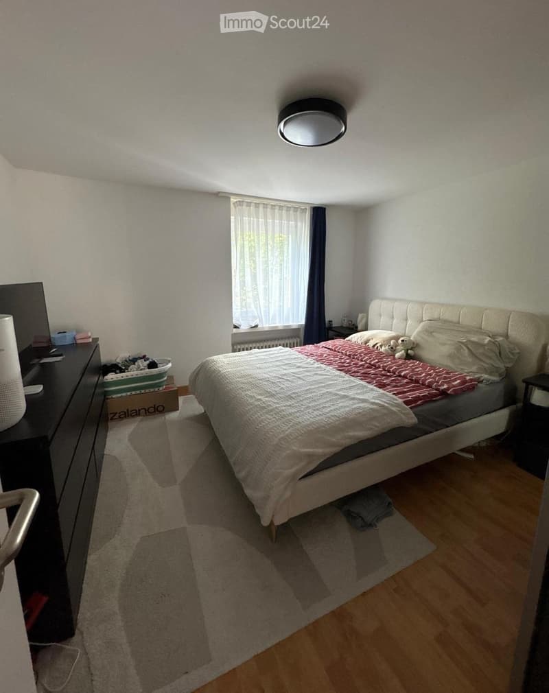 Bedroom with a large bed, nightstand, lamp, and a window with curtains. The room features wooden flooring and a rug.