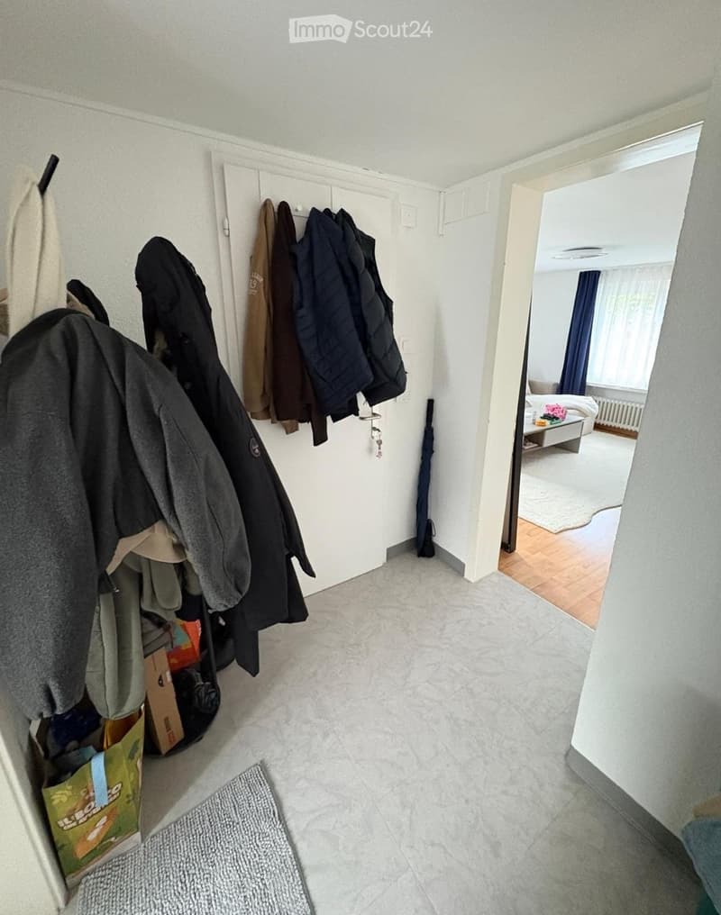 A clean, well-lit hallway with tiled flooring, white walls, and a coat rack holding multiple jackets. An umbrella is placed near the coat rack, and a small rug lies on the floor. An open doorway leads to a bedroom with a bed, nightstand, and window with c