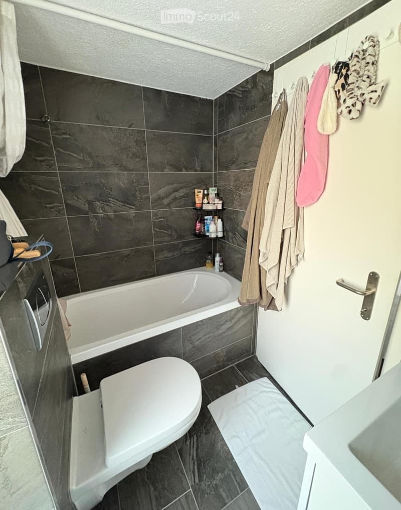 Modern bathroom with dark grey tiles, a bathtub, toilet, towel rack, and a shelf with toiletries.