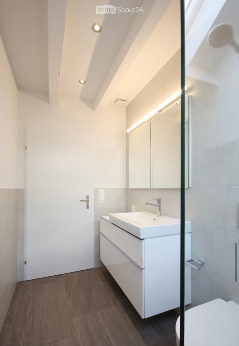 Modern bathroom with a white sink, chrome faucet, built-in cabinet, and a glass-enclosed shower area. The room features wooden flooring and recessed lighting.