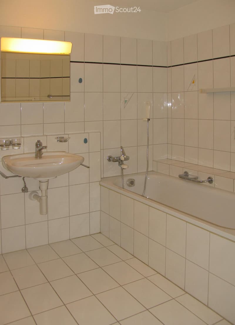 Bathroom with white tiled walls and floor, featuring a sink with a mirror above it, a bathtub with a handheld showerhead, and a built-in shelf on the wall.