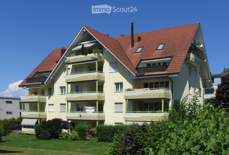 A two-story house with a red-tiled roof, multiple balconies, and green shutters. The house is surrounded by a well-maintained garden with various plants and shrubs.