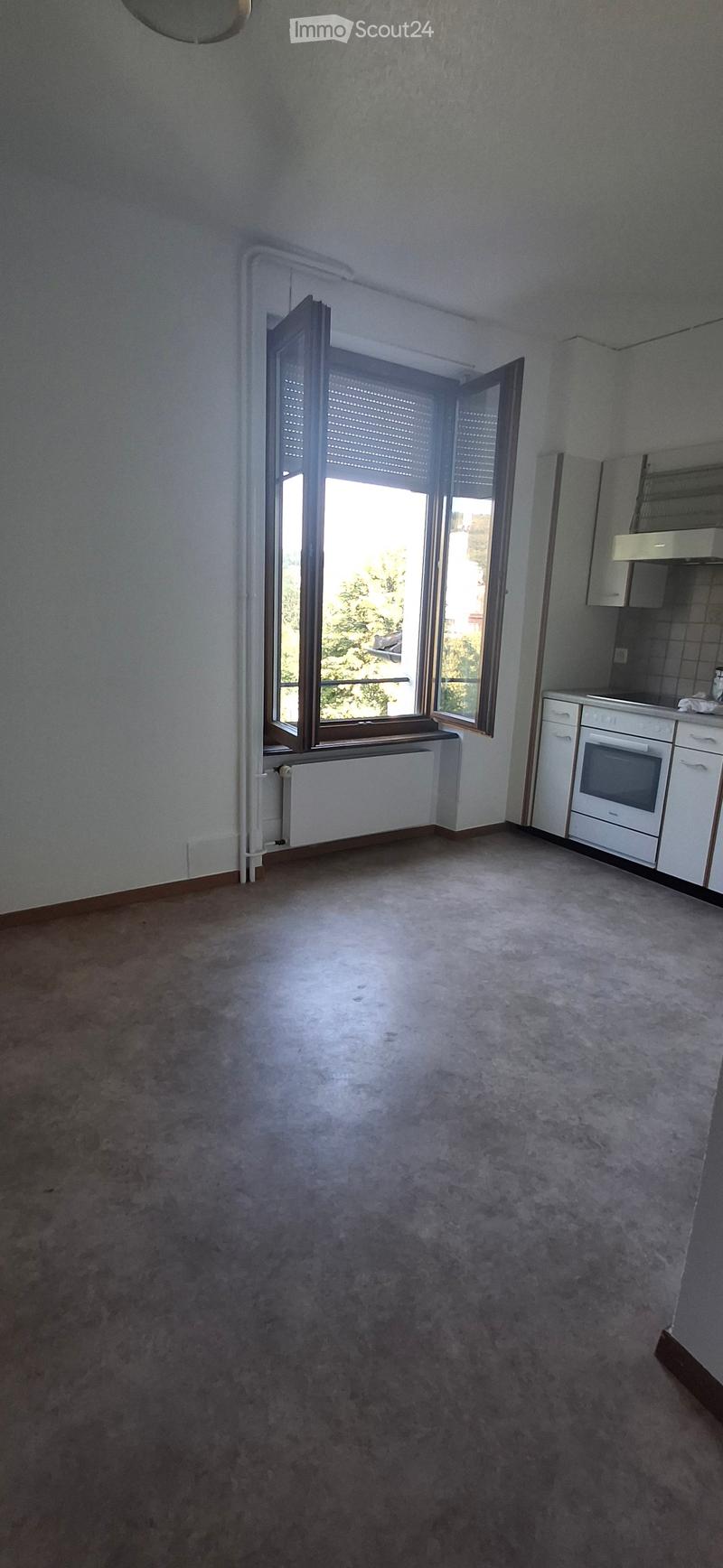 Empty room with carpeted floor, white walls, large window with shutters, radiator, and small kitchen area with cabinets and oven.