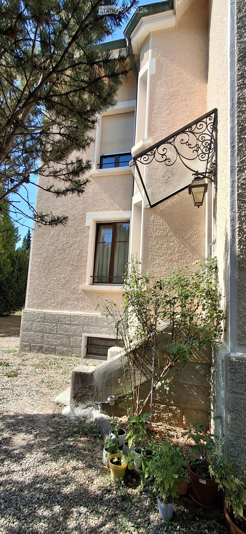Two-story house with beige stucco exterior, multiple windows, decorative wrought-iron balcony, and small garden area with potted plants and gravel ground.