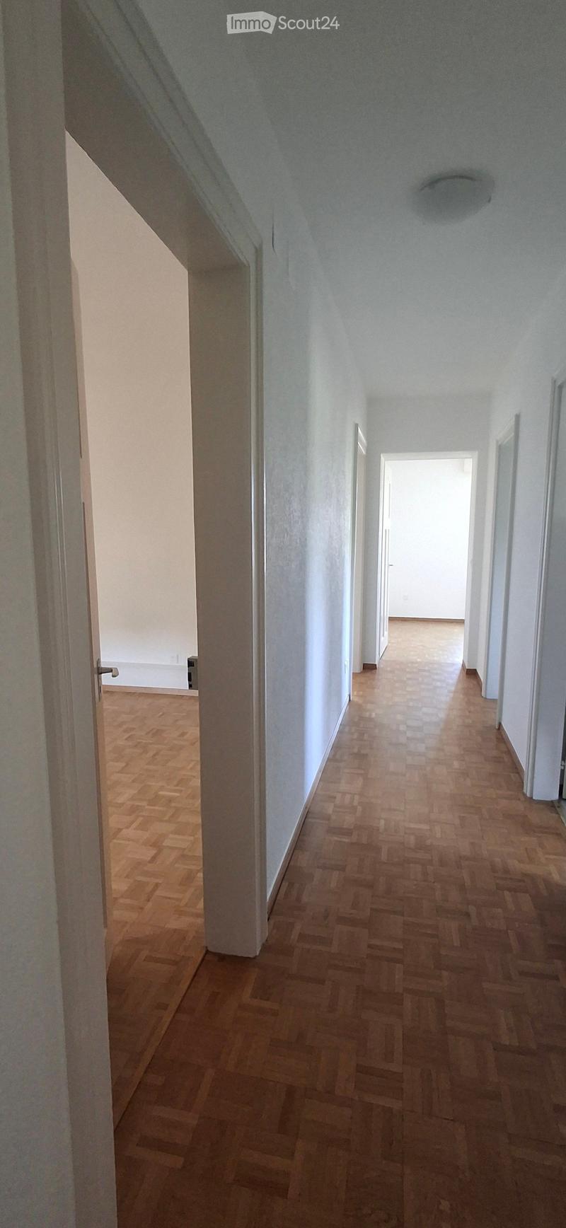 Empty hallway with wooden flooring and white walls, featuring a visible ceiling light fixture and an open doorway at the end.