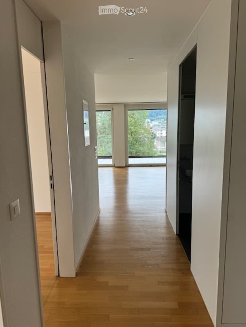Bright hallway with wooden flooring, white walls, large windows, and a view of greenery outside.