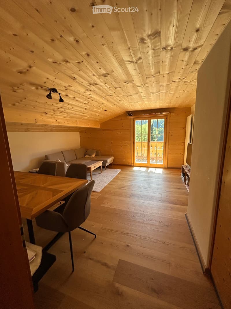 Bright, wooden-floored room with exposed wooden ceiling beams, a sofa, coffee table, ottoman, and a dining area with a table and chairs.