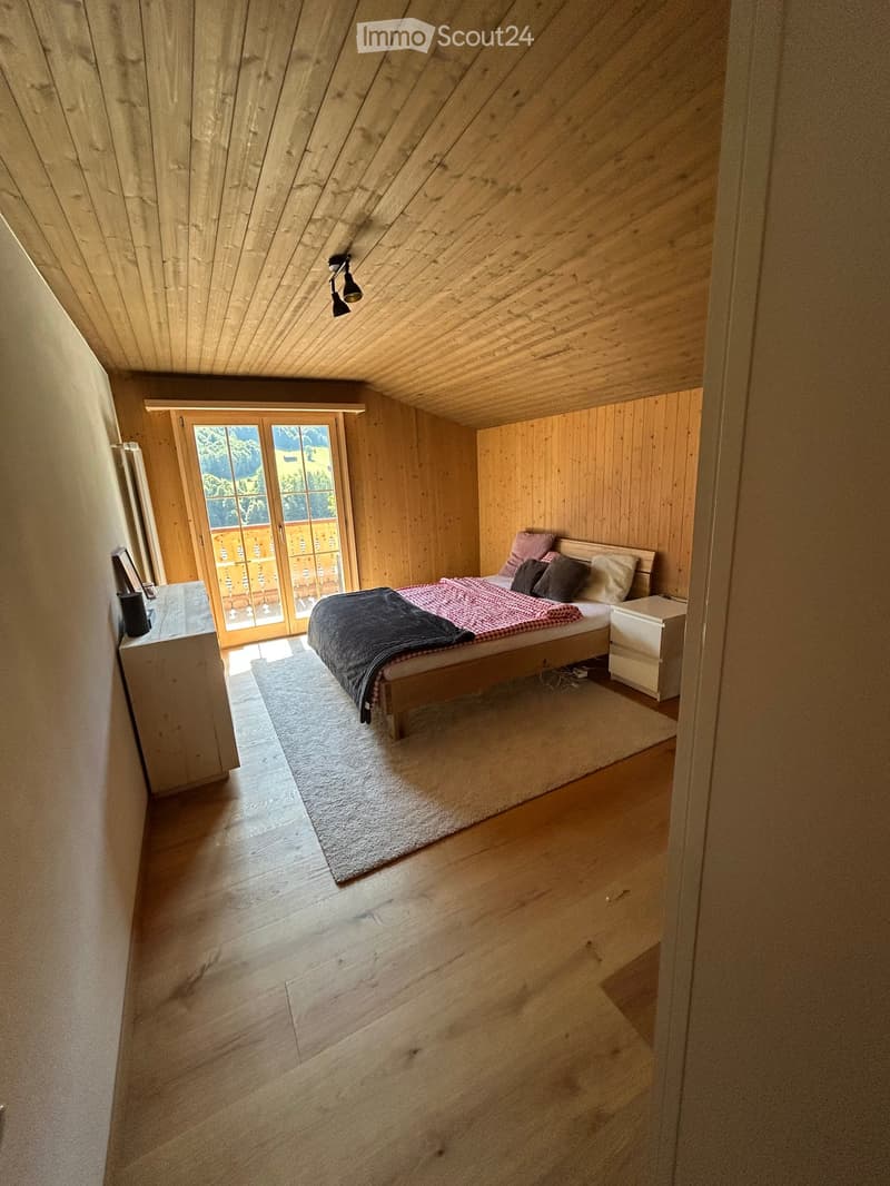 Cozy attic bedroom with wooden walls and ceiling, large window with view, bed with pillows and blankets, nightstand, and lamp.