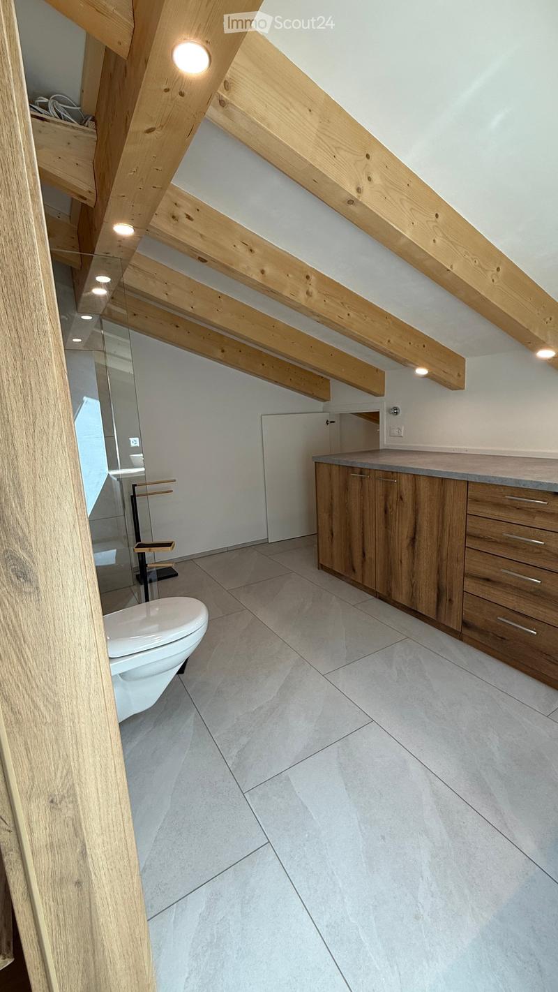 Modern bathroom with exposed wooden beams, white tiled floor, and a white toilet. Features a wooden cabinet with drawers and a countertop, and a glass-enclosed shower area visible in the background.