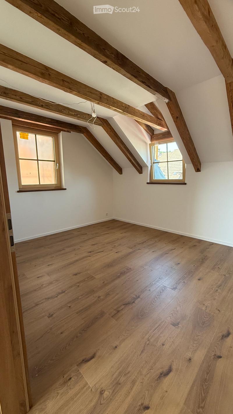 Empty attic room with wooden beams, wooden flooring, and two windows, one with frosted glass.