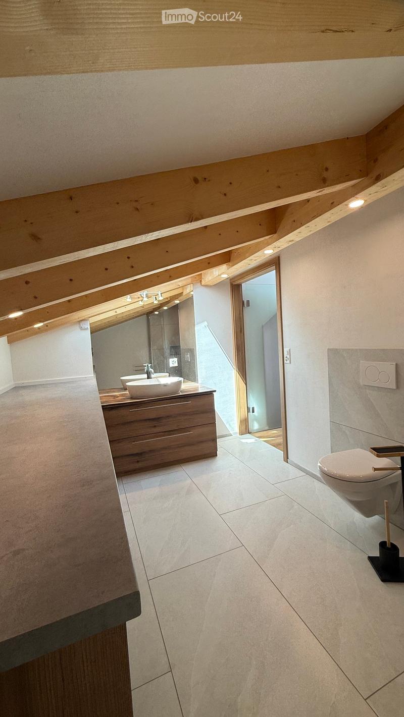 Modern bathroom with wooden ceiling beams, white tiled floor, double sink vanity with wooden cabinet, toilet, and a glass-enclosed shower area visible through an open doorway.