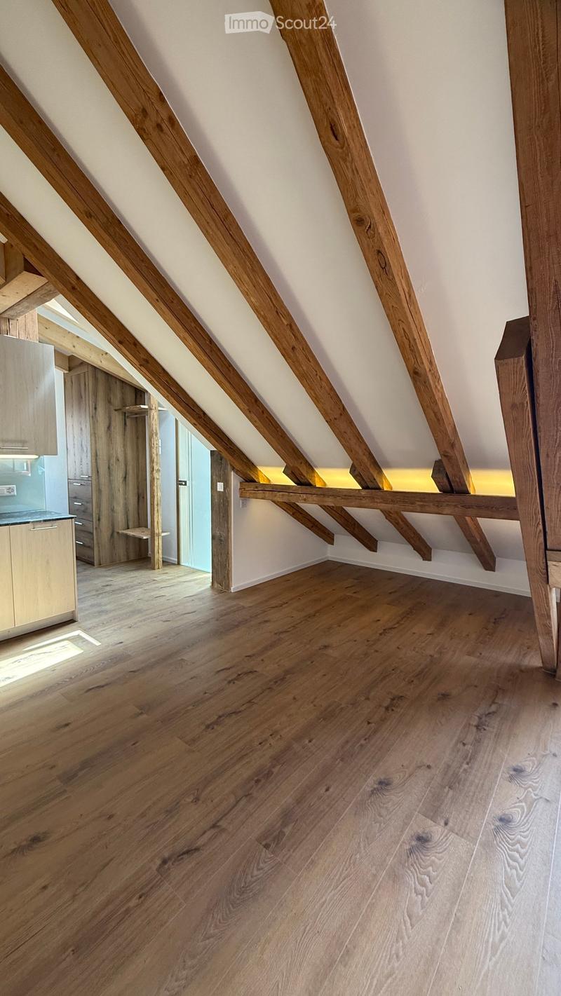 An attic space with exposed wooden beams and a wooden floor, featuring a small built-in shelf and a doorway leading to another room.
