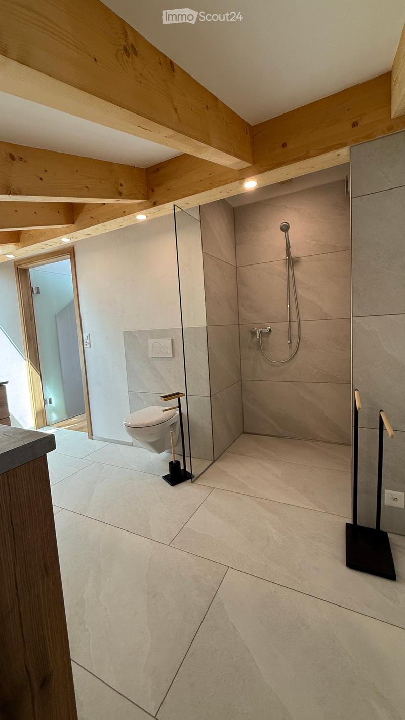 Modern bathroom with light-colored tiles, a glass-enclosed shower area, a toilet, and a sleek shower hose. The ceiling features exposed wooden beams and recessed lighting.