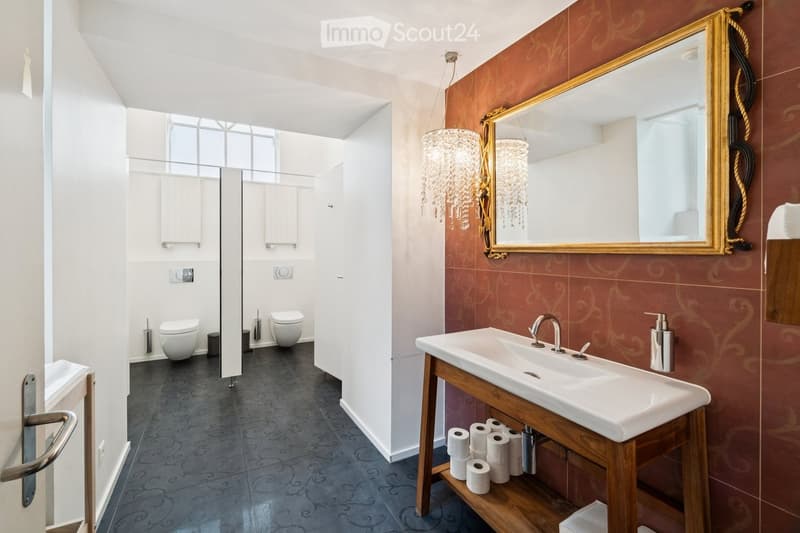 Modern bathroom with a double sink, ornate mirror, chandelier, and two toilets in a separate compartment.