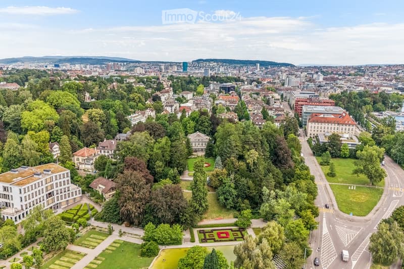 Aerial view of a cityscape with a mix of residential and commercial buildings, surrounded by green spaces and trees. The area includes a park with a pond, pathways, and a patterned garden bed.