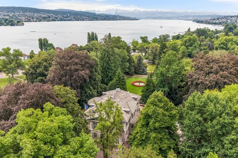 A large, multi-story house surrounded by lush greenery, situated near a lake with distant mountains and a cityscape visible in the background.