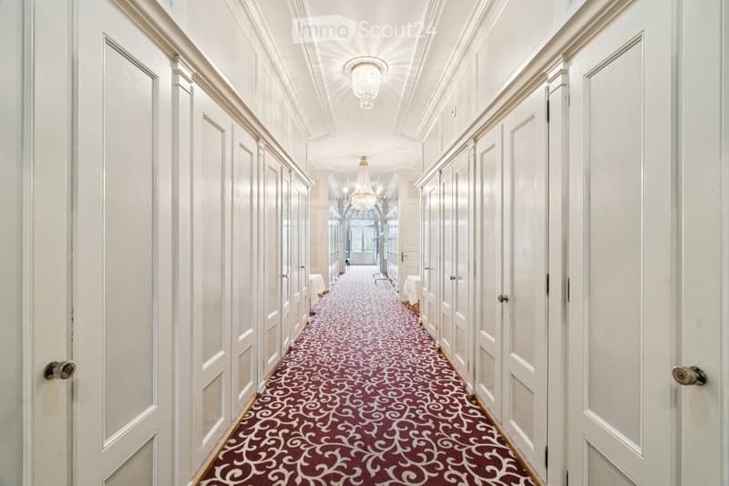 Elegant hallway with white paneled walls, ornate carpet, chandeliers, and multiple doors.
