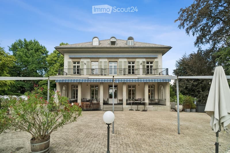 A two-story building with a balcony and outdoor seating area, surrounded by greenery and a paved courtyard.