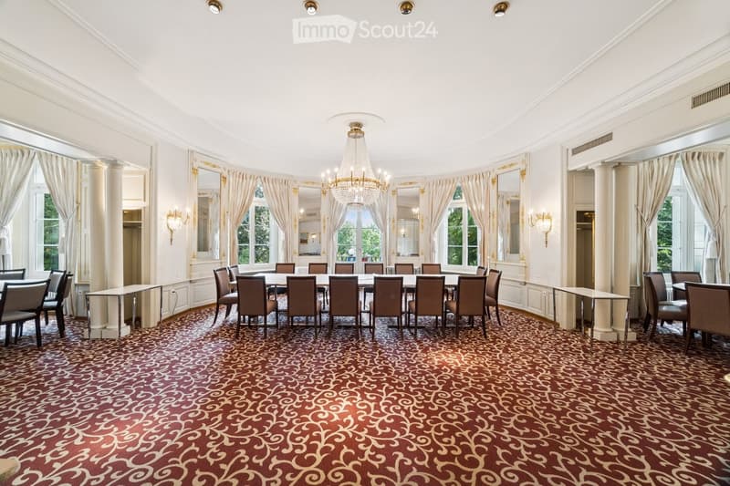 Elegant dining room with a large round table surrounded by chairs, a chandelier hanging from the ceiling, side tables, large windows with curtains, and a patterned carpet.